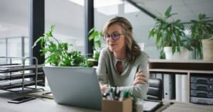 Laptop, arms crossed and mature business woman at desk in glass office for administration or online report. Computer, email and research with confident employee at table in professional workplace